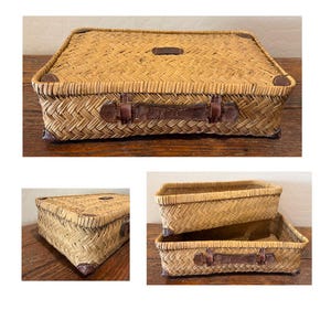 May include: Woven wicker storage basket with a rectangular shape and a lid. The basket features brown leather accents on the corners, handle, and lid. The basket is made of light brown wicker.