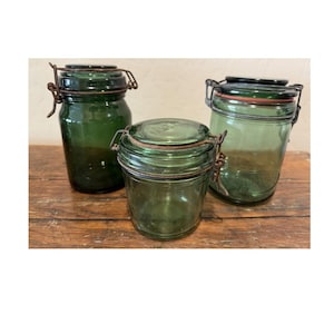 May include: Three green glass jars with metal closures and hinged lids. The jars are different sizes and shapes, with a vintage aesthetic. The jars are displayed on a wooden surface.