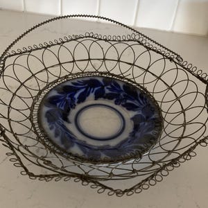 May include: An antique wire basket with a handle, holding a blue and white patterned plate. The basket has a geometric design and a decorative edge. The plate features a floral design.