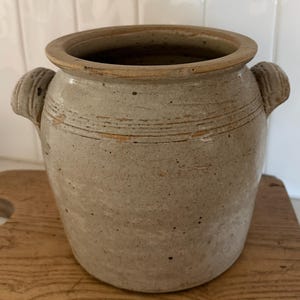 May include: A light beige stoneware crock featuring a wide circular opening, a thick rim, and two small side handles. The exterior surface displays a speckled texture with horizontal incised lines around the upper section and visible wear.