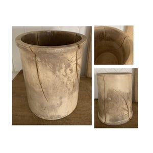 May include: A cylindrical, antique stoneware crock with a wide opening and a thick rim. The off-white, aged surface shows visible cracks and a weathered appearance, suggesting its age and use. The crock sits on a wooden surface.