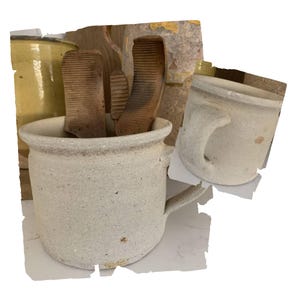May include: A rustic, off-white ceramic mug with a handle, holding several wooden washboards. The mug has a textured surface and a slightly flared rim. Another similar mug is visible to the right. The background includes a yellow ceramic jar and a weathered wall.