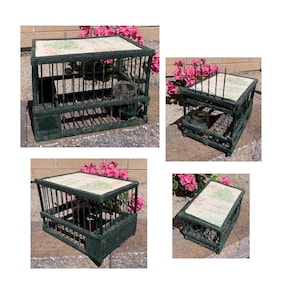 May include: A dark green decorative birdcage with a rectangular shape. The cage has vertical bars and a patterned top with a floral design. A small metal container is inside the cage. The cage is outdoors near pink flowers.
