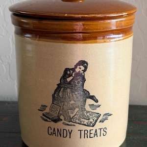 May include: A cylindrical ceramic canister with a glossy brown lid and a tan body. The front features a black graphic illustration of a person sitting on a stool with a book and the text CANDY TREATS printed in a simple sans-serif font below.