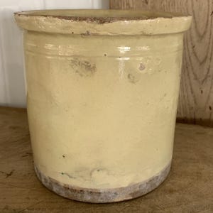May include: A cylindrical, antique-style ceramic jar with a light yellow glaze. The jar has a slightly flared rim and a rough, aged appearance, with visible imperfections and a darker base. The overall aesthetic suggests a vintage or rustic design.