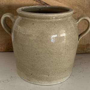 May include: A beige ceramic pot with two curved handles. The pot has a wide opening and a slightly textured surface. Horizontal lines encircle the pot near the top. The pot is likely used for storage or decoration.