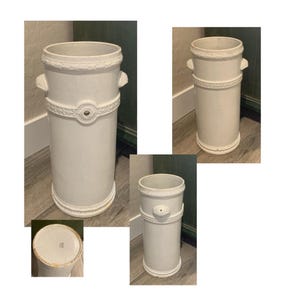 May include: A white ceramic umbrella stand with a cylindrical shape. It features decorative rope-like trim around the top and middle, with small handles and a central medallion. The base has a similar rope-like trim.