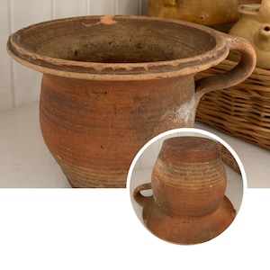 May include: A rustic, terracotta-colored pottery vessel with a wide, shallow opening and a curved handle. The aged pot has a rounded body and a circular base. The image also includes a smaller, inverted version of the pot, showcasing its shape.