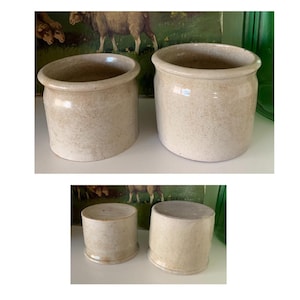 May include: A collection of four antique stoneware crocks in varying sizes. The crocks are a speckled beige color with a rounded shape and a wide opening. The crocks are displayed on a white surface with a green background.