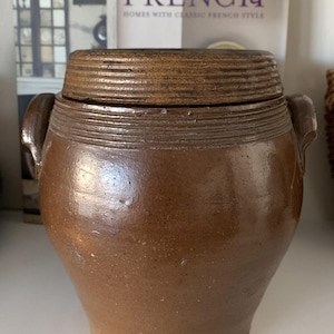 May include: A large, brown stoneware crock with a lid. The crock has two handles and a textured surface. The lid has a series of ridges around the top.