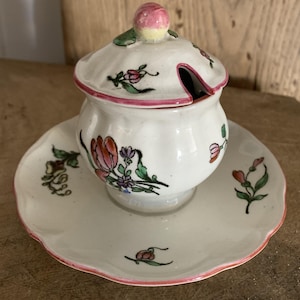 May include: A white ceramic sugar bowl with a lid and saucer, adorned with floral designs in pink, green, and purple. The rim of the saucer and bowl is outlined in pink. The lid features a pink and yellow knob.