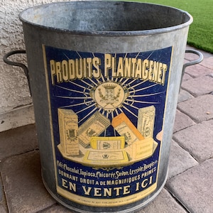 May include: A galvanized metal bucket with two handles and a blue and yellow label. The label reads "PRODUITS PLANTAGENET" and features illustrations of various products. The text "EN VENTE ICI" is also visible.