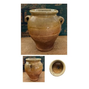 May include: A tan and brown glazed ceramic pot with two small side handles and a wide, rounded rim. The vessel features a tapered body with a smooth, glossy finish and a circular opening at the top, showing a hollow interior and a flat base.