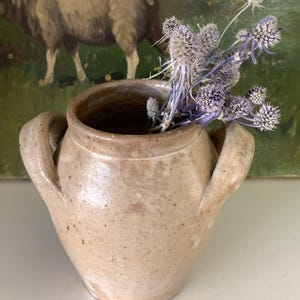 May include: A small, beige ceramic vase with two handles, filled with dried purple flowers. The vase has a speckled texture and a rustic appearance. The background features a painting of a sheep.