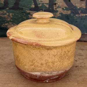May include: A small, round, lidded ceramic jar with a glossy mustard-yellow glaze. The vessel features a smooth, domed lid with a central knob handle. The base of the jar reveals a contrasting reddish-brown clay rim, showing a weathered texture.