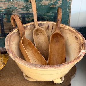 May include: A large, round, light yellow ceramic bowl with a wide rim and a footed base, containing three individual wooden scoops. Each scoop features a long, curved handle and a deep, elongated basin, showing natural wood grain and texture.