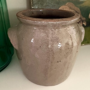 May include: A vintage, gray-brown stoneware crock with two side handles. The ceramic pot has a wide opening and a slightly darker rim. The surface shows a crackled texture, suggesting age and use. The crock is likely for kitchen or home decor.