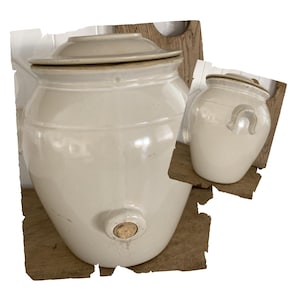 May include: Off-white ceramic crock with a lid, featuring a spigot and a cork. A smaller matching crock with a handle and a decorative design is also present. Both are on a wooden surface, showcasing a rustic aesthetic.