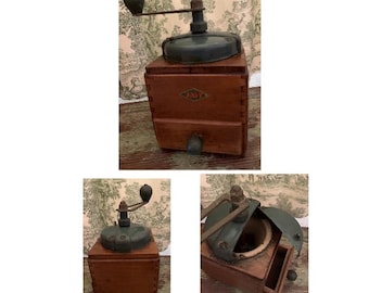 May include: A square wooden coffee grinder featuring a dark metal hand crank, a green metal lid, and a small pull-out drawer. The front of the wooden box displays a diamond-shaped logo with the text JURA in red and black lettering.