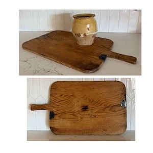 May include: A rectangular wooden cutting board with a handle and dark metal corner accents, featuring a prominent wood grain pattern. A small, round, tan-colored ceramic pot with a wide rim sits on the board in the top image view.