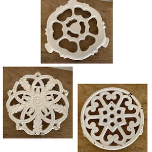 May include: Three white cast iron trivets with decorative designs. One has a floral pattern, another has a geometric design, and the third has a unique openwork design. They are displayed on a wooden surface.