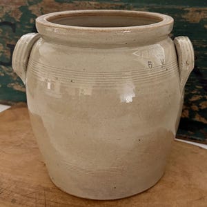 May include: A light beige stoneware crock featuring a wide mouth, a thick rolled rim, and two side handles. The exterior surface displays horizontal incised lines around the upper and lower sections, with the number 5 stamped on the upper body.