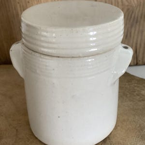 May include: A cream-colored ceramic jar with a lid. The jar has a cylindrical body with two small handles and a lid with horizontal ridges. The jar is sitting on a wooden surface.