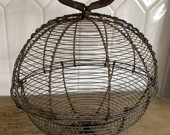 May include: A vintage wire basket with a round, cage-like design. The basket has a circular handle at the top and sits on a small, circular base. The metal has a rustic, aged appearance, with visible signs of wear and oxidation.