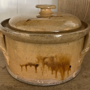 May include: A tan ceramic lidded pot with two side handles. The pot has a round shape with a lid and a small knob on top. The pot has dark brown drips down the side.