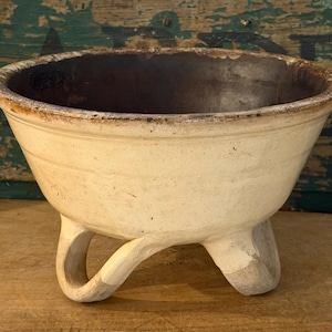 May include: A cream-colored ceramic bowl with a wide, flared rim and a dark brown interior. The bowl features three curved, sturdy legs that elevate the base. The exterior surface shows signs of wear with visible texture and discoloration.