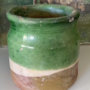 May include: A small, antique ceramic pot with a rounded body and a slightly flared rim. The pot is painted in three horizontal bands of color: green, white, and brown. The green band is at the top, the white in the middle, and the brown at the bottom. The pot has a rustic, aged appearance.
