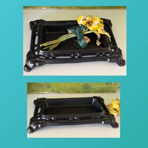 May include: A rectangular black metal tray with an ornate, openwork border and four decorative feet. The tray features a smooth, recessed center. In one view, a small bouquet of yellow and white artificial flowers with a green ribbon rests inside.