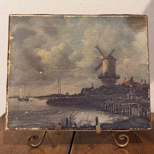 May include: A rectangular painting on canvas featuring a landscape scene with a prominent windmill, a body of water, and a sailboat. The color palette consists of muted blues, grays, and browns, depicting a cloudy sky over a coastal village setting.