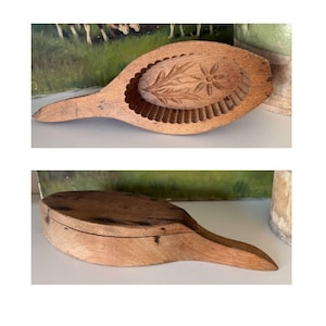 May include: A vintage wooden butter mold with a handle. The mold features a carved floral design within a rectangular frame. The second image shows the mold closed. The wood has a warm, natural tone.