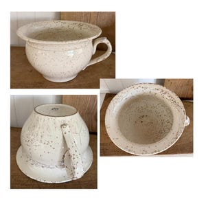 May include: Off-white ceramic chamber pot with a handle and a wide rim. The pot has a distressed, speckled finish with visible cracks, giving it a vintage appearance. The interior and exterior show signs of wear, enhancing its antique aesthetic.