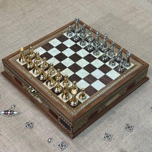 May include: A wooden chess set with a checkerboard pattern. The chess pieces are gold and silver. The board is inlaid with mother-of-pearl and dark wood. The set is displayed on a tan surface.