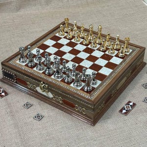 Puede incluir: Un juego de ajedrez de madera con un tablero a cuadros marrón y nácar. Las piezas del ajedrez son doradas y plateadas. El juego está en una caja de madera con incrustaciones decorativas y un cierre. Varias baldosas decorativas están esparcidas alrededor del juego.