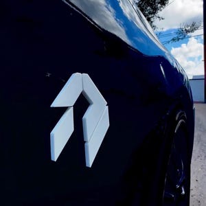 May include: A glossy black car door panel featuring a white, angular, geometric emblem consisting of three distinct, sharp-edged segments arranged in a vertical, arrow-like formation. The surface reflects the bright blue sky and white clouds.