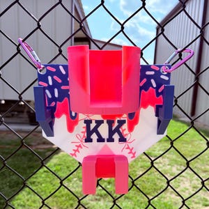 May include: A baseball-themed phone holder in white, blue, and pink. The holder displays the initials "KK" and a baseball stitch design. It is attached to a chain-link fence using pink carabiner clips.