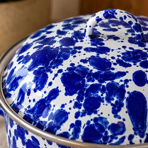 May include: A round cooking pot with a matching lid, featuring a white base with a vibrant blue speckled pattern. The pot has a metallic silver rim and a curved handle on the lid, showcasing a glossy finish throughout the entire exterior surface.