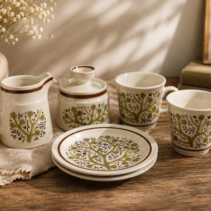 May include: A ceramic tea set featuring a cream-colored base with a repeating olive green tree motif and brown rim accents. The set includes a creamer pitcher, a lidded sugar bowl, two mugs, and two small saucers, all decorated with the same pattern.