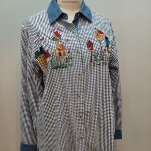 May include: A blue and white gingham button-down shirt with a denim collar and cuffs. The shirt features embroidered birdhouses and floral accents. The text on the shirt reads "Live Well, Laugh Often, Love Much."