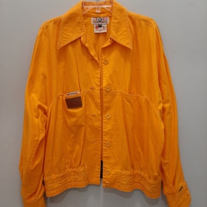 May include: A bright yellow, cropped jacket with a button-up front and a single pocket on the left side. The jacket has a gathered elastic waistband and long sleeves with buttoned cuffs.