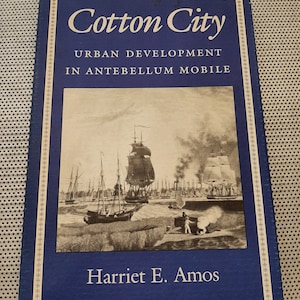 May include: A blue book with a gold border and the title "Cotton City: Urban Development in Antebellum Mobile" by Harriet E. Amos. The cover features a black and white photograph of a harbor scene with ships and a man standing on a dock.
