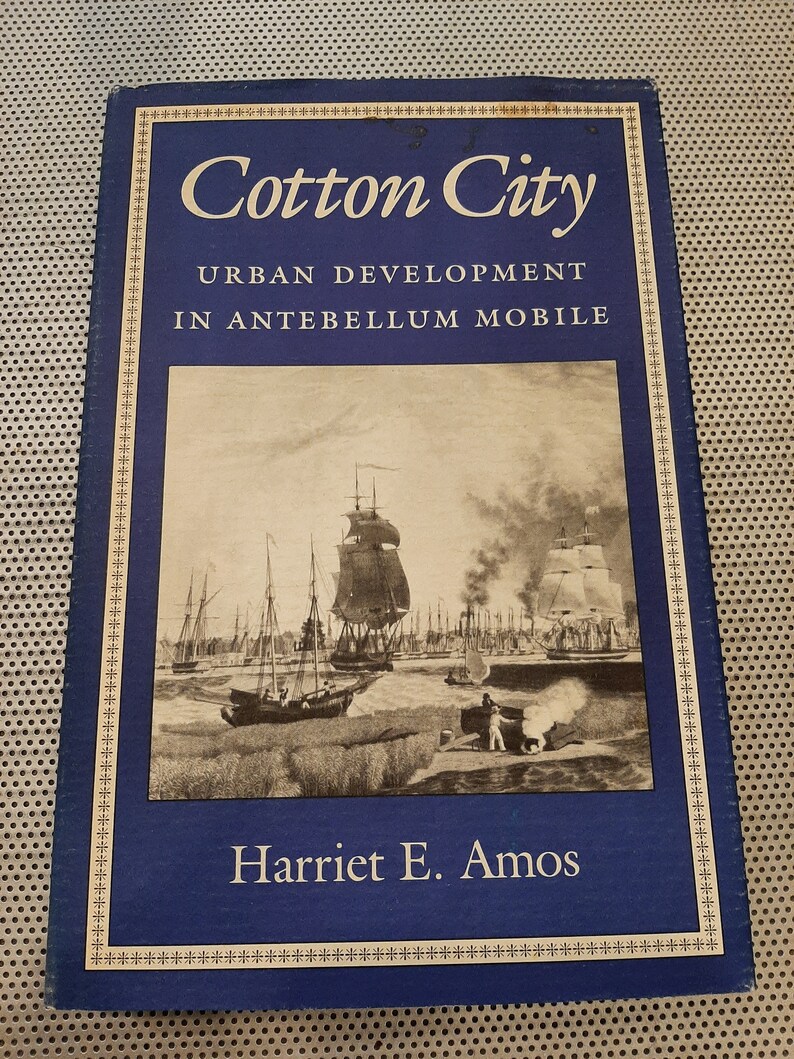 Peut inclure: Un livre bleu avec une bordure dor&eacute;e et le titre "Cotton City: Urban Development in Antebellum Mobile" par Harriet E. Amos. La couverture pr&eacute;sente une photographie en noir et blanc d'une sc&egrave;ne de port avec des navires et un homme debout sur un quai.