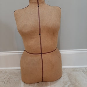 May include: A vintage, beige fabric dress form with brown tape markings. The form is missing a head and has a rounded torso and hips.