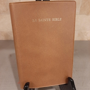 May include: A tan-colored Bible with a smooth, leather-like cover. The words "LA SAINTE BIBLE" are embossed in gold lettering on the front cover. The book is held upright by a black plastic display stand, showcasing its rectangular shape.