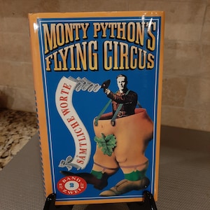 May include: A book titled "Monty Python's Flying Circus" with a blue cover and orange border. The cover features an illustration of a man in a pair of pants. The book is propped up on a black stand.