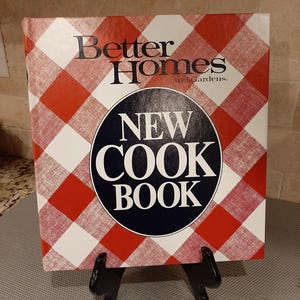May include: A cookbook titled "Better Homes and Gardens New Cook Book" with a red and white checkered pattern cover. The title is in black and the words "New Cook Book" are in a navy blue circle. The book is displayed on a black stand.