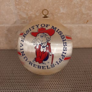 May include: A round Christmas ornament with the words "UNIVERSITY OF MISSISSIPPI REBELS" encircling a cartoon figure in a red hat and suit. The ornament has a gold-colored metal loop at the top for hanging.